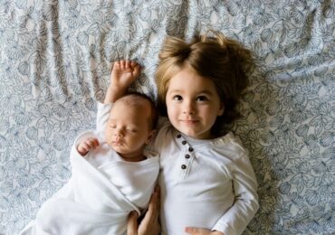 : a small girl and a baby lying on a bed symbolizing things to consider when buying a house with kids