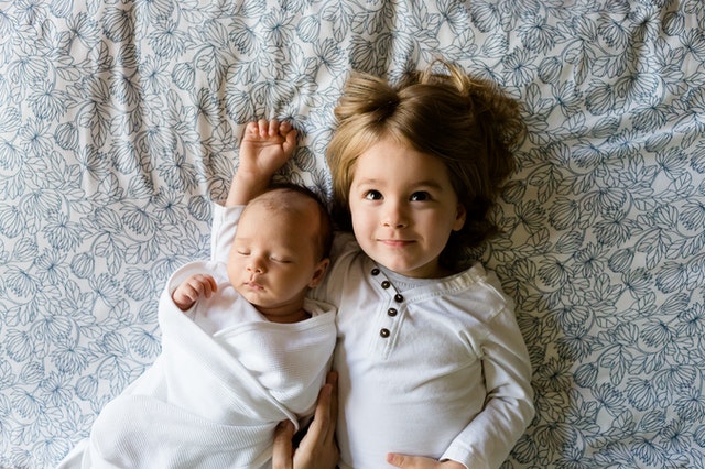 : a small girl and a baby lying on a bed symbolizing things to consider when buying a house with kids