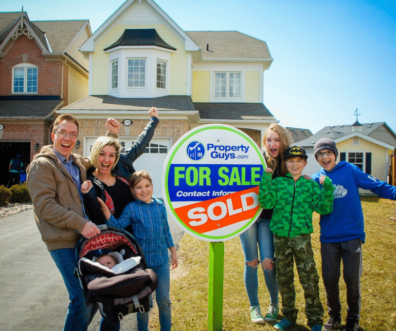 Family happily celebrating the sale of their home in front of a PropertyGuys.com “Sold” sign.