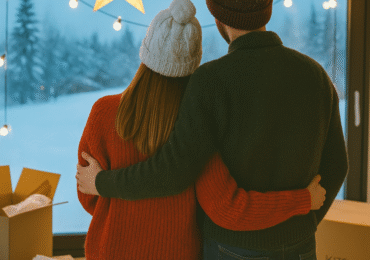 A couple dressed warmly in winter sweaters and hats stands arm in arm by a large window, looking out at a snowy landscape with moving boxes nearby, symbolizing a winter move.