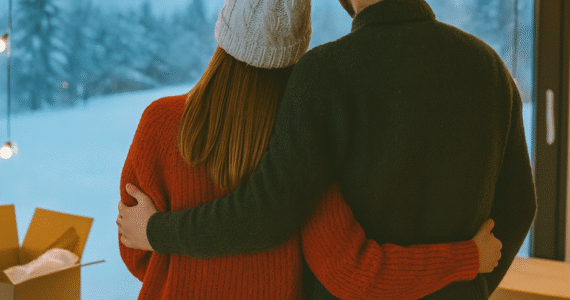 A couple dressed warmly in winter sweaters and hats stands arm in arm by a large window, looking out at a snowy landscape with moving boxes nearby, symbolizing a winter move.