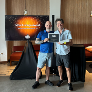 Two men standing indoors holding a framed "Most Listings Award." In the background, a television screen displays the "Rewy Awards" logo and "Most Listings Award: Acadie, Chaleur & Restigouche, NB" against a sparkling orange and black backdrop.