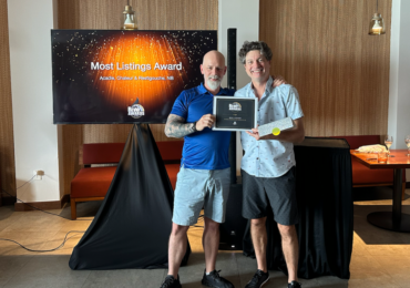 Two men standing indoors holding a framed "Most Listings Award." In the background, a television screen displays the "Rewy Awards" logo and "Most Listings Award: Acadie, Chaleur & Restigouche, NB" against a sparkling orange and black backdrop.