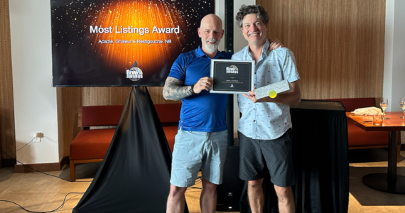 Two men standing indoors holding a framed "Most Listings Award." In the background, a television screen displays the "Rewy Awards" logo and "Most Listings Award: Acadie, Chaleur & Restigouche, NB" against a sparkling orange and black backdrop.