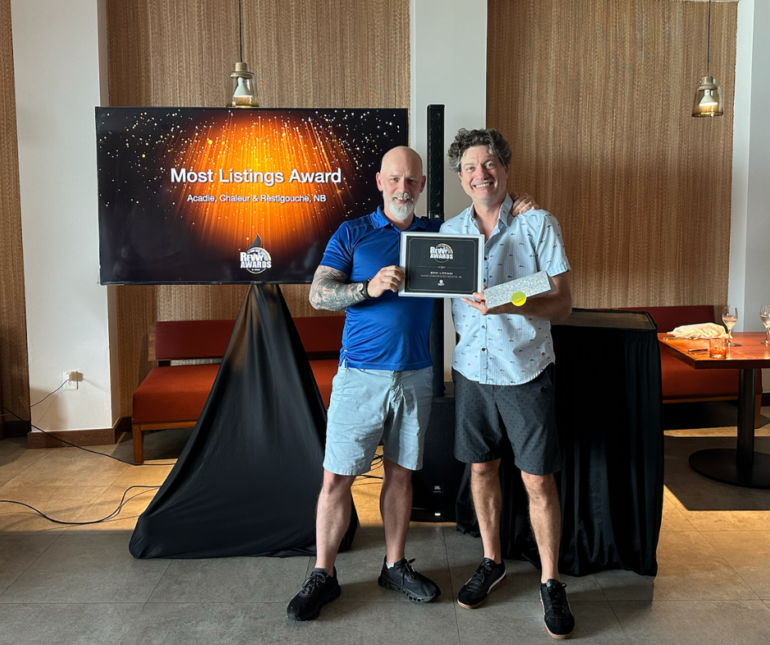 Two men standing indoors holding a framed "Most Listings Award." In the background, a television screen displays the "Rewy Awards" logo and "Most Listings Award: Acadie, Chaleur & Restigouche, NB" against a sparkling orange and black backdrop.