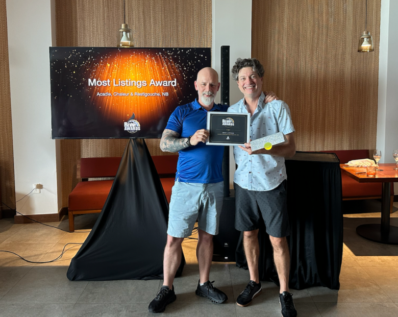 Two men standing indoors holding a framed "Most Listings Award." In the background, a television screen displays the "Rewy Awards" logo and "Most Listings Award: Acadie, Chaleur & Restigouche, NB" against a sparkling orange and black backdrop.